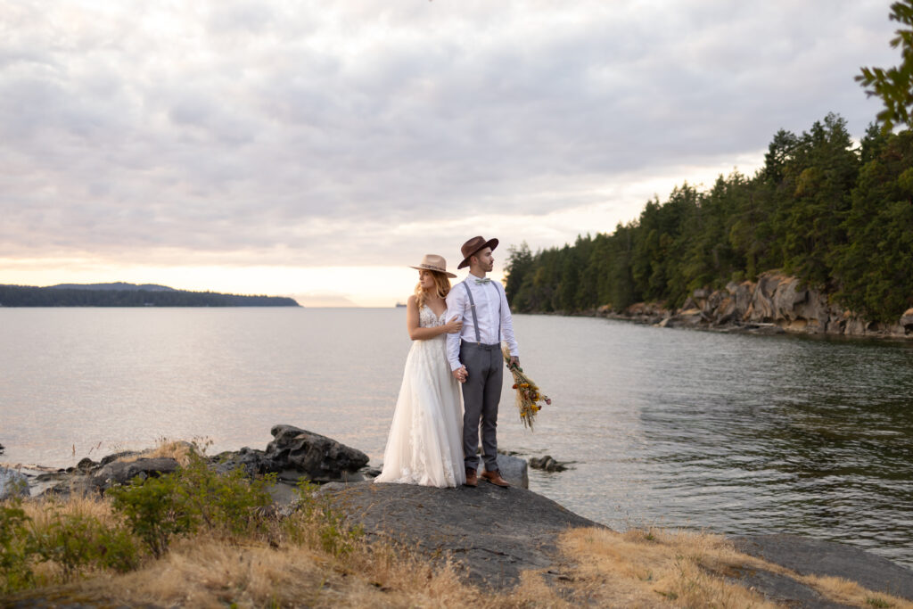 Couple stands next to ocean wearing wedding attire and wide brimmed hats